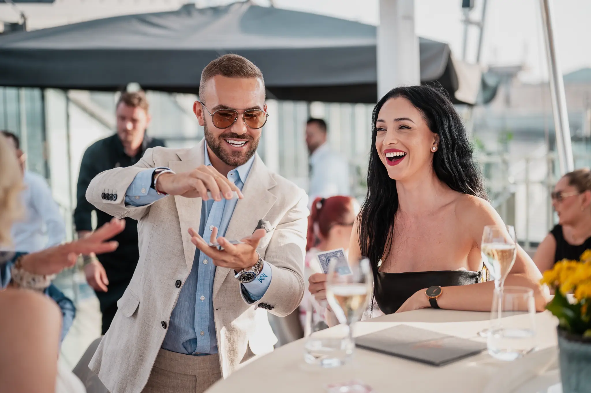 Matteo Cammisa performing close-up magic at an outdoor cocktail terrace with champagne glasses and guests laughing
