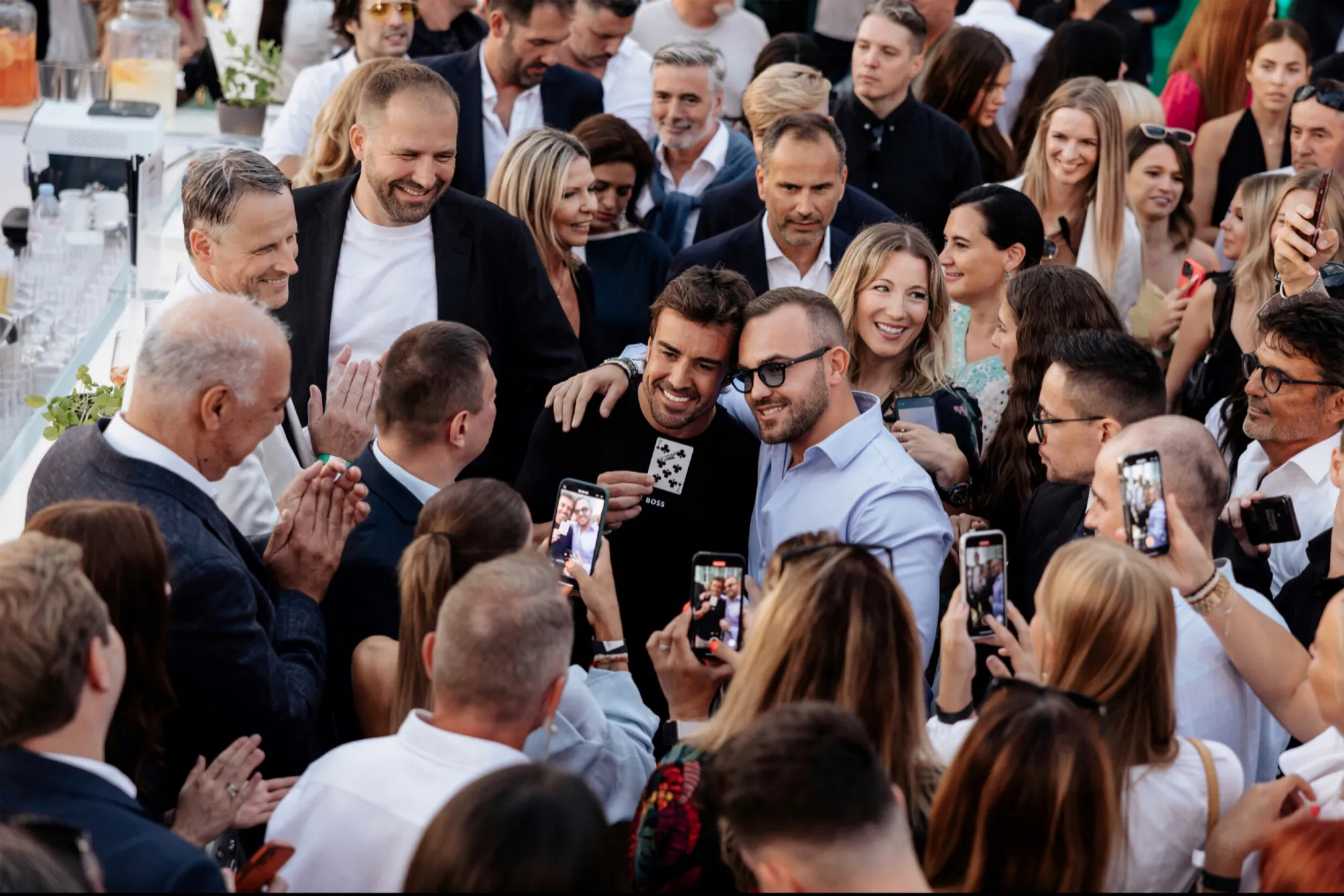 Fernando Alonso holds up a signed playing card beside magician Matteo Cammisa, surrounded by a crowd photographing the moment