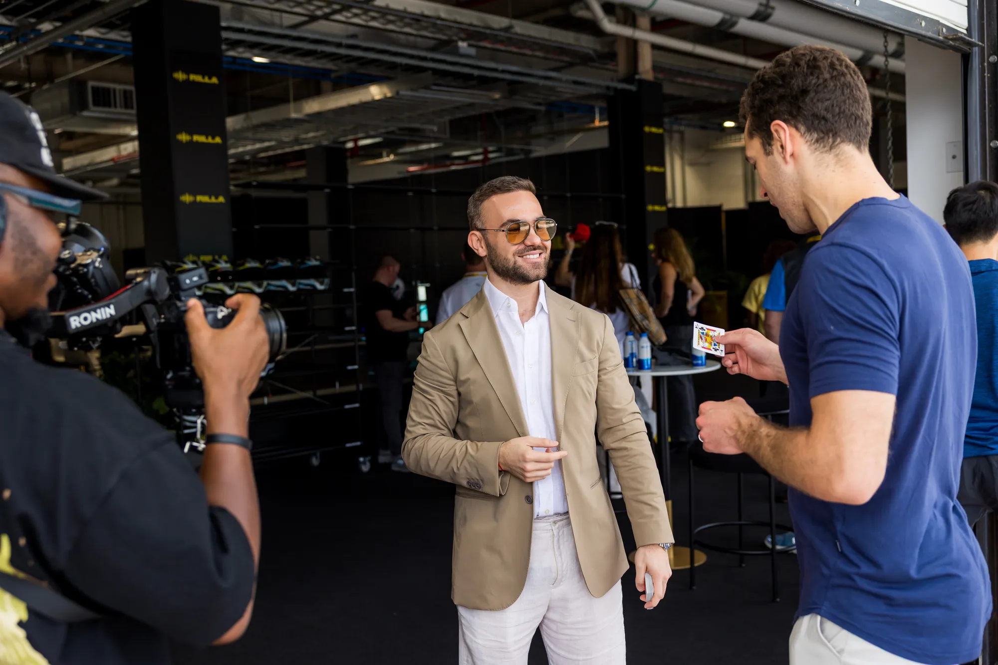 Matteo Cammisa performing close-up card magic for a guest in a race-week hospitality space while a videographer films