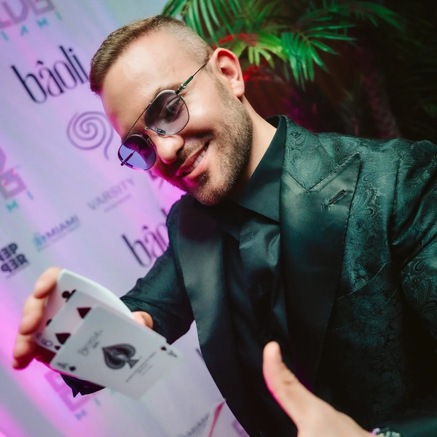 Matteo Cammisa in a dark paisley dinner jacket fanning playing cards on the Baoli Miami step-and-repeat wall at The GR1D Club's Formula 1 afterparty