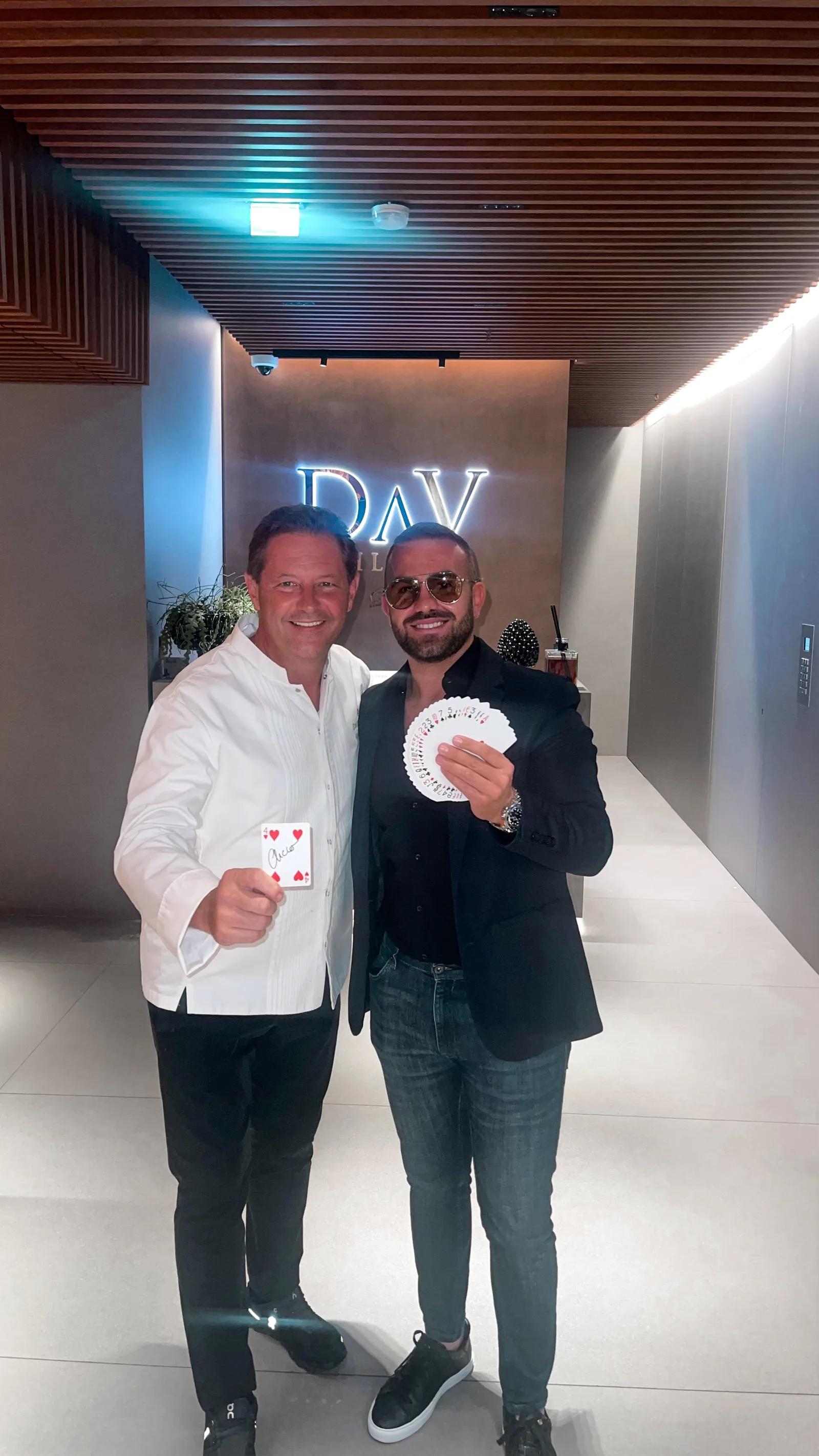 A chef in whites holding a signed four of hearts beside Matteo Cammisa, who fans a deck of cards, under the illuminated DaV sign in Milan