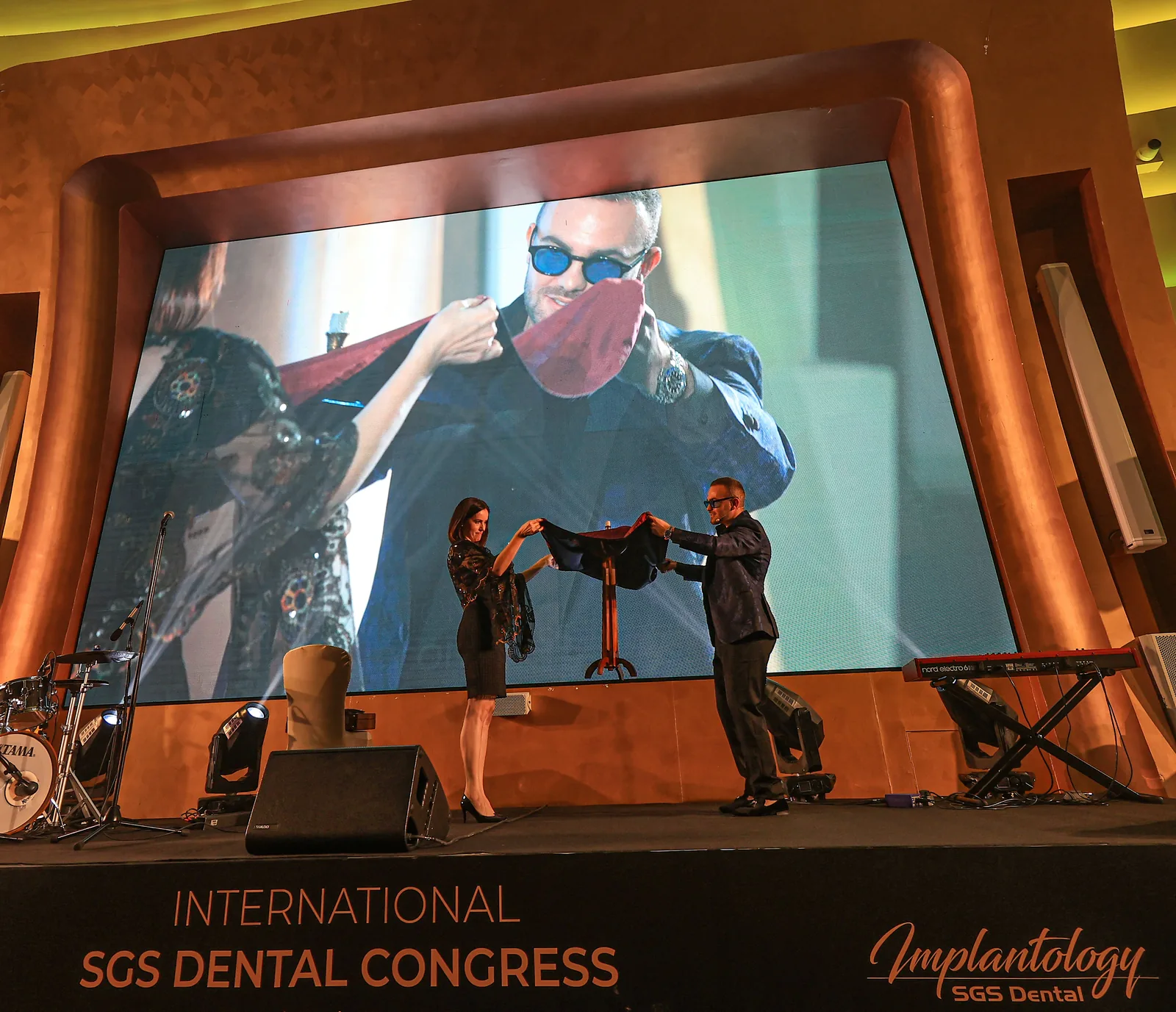 Matteo Cammisa on stage with a volunteer beneath the International SGS Dental Congress banner at the V Hotel Dubai, the effect shown live on the screen behind them