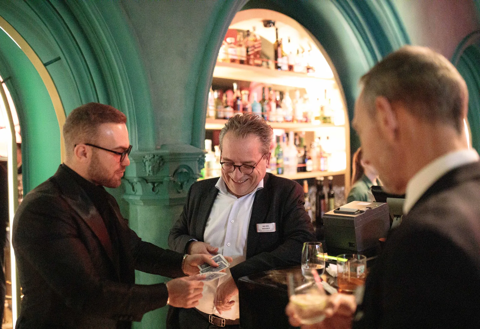 Matteo Cammisa performing a card effect at the bar of W Budapest, guests smiling and leaning in beneath the hotel's deep green arches